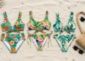Folded tropical-print bikini sets arranged on sand with a beach bag