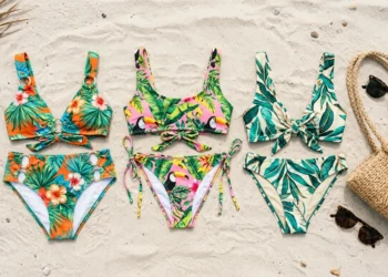 Folded tropical-print bikini sets arranged on sand with a beach bag