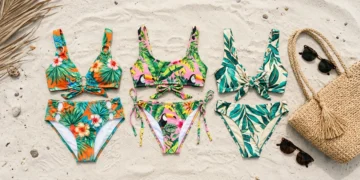 Folded tropical-print bikini sets arranged on sand with a beach bag