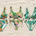 Folded tropical-print bikini sets arranged on sand with a beach bag