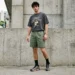 A young man wearing a washed dark grey graphic T-shirt, muted olive green casual shorts, black crew socks, and stylish sneakers, standing in front of a concrete wall in a modern urban streetwear setting.